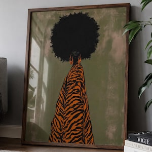 May include: A framed wall art print featuring a stylized illustration of a person with a large black afro hairstyle and orange earrings, wearing a flowing orange and black tiger-striped garment against a muted olive green background.