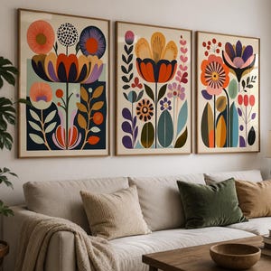Può includere: Un set di tre stampe artistiche da parete incorniciate con illustrazioni floreali stilizzate in vivaci tonalità di arancione, viola, blu e giallo. Ogni stampa rettangolare mostra motivi botanici simmetrici con petali, foglie e steli.