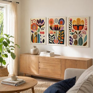 Three framed colorful folk floral wall art prints displayed above a cream sofa in a modern living room with warm wood frames, orange pillows, patterned throw, coffee table, and indoor plant.