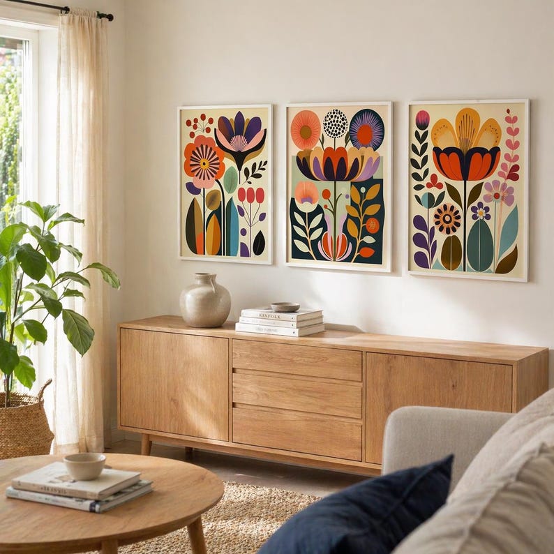 Three framed colorful folk floral wall art prints displayed above a cream sofa in a modern living room with warm wood frames, orange pillows, patterned throw, coffee table, and indoor plant.