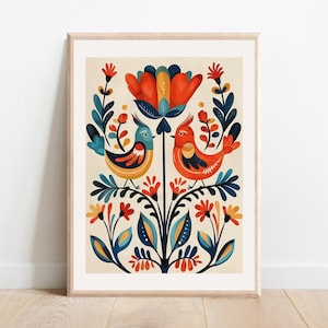 A framed art print with a symmetrical design of two birds perched on a floral stem. The birds are orange and blue, with a large orange and blue flower above them. The artwork has a folk art style with a cream background.