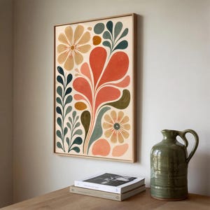 May include: A framed floral art print featuring a vibrant design with orange, peach, and green abstract flowers and leaves. The artwork is displayed on a light-colored background, framed in a natural wood frame. A green ceramic pitcher and books are on a wooden table.