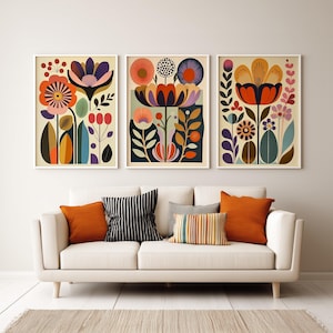 Framed colorful folk floral wall art print hanging above a wood table in a minimal neutral room, styled with stacked books and a green ceramic pitcher.