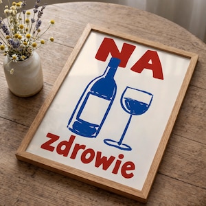 Puede incluir: Un cuadro enmarcado con una ilustración azul de una botella y una copa de vino, con el texto rojo "NA" arriba y "zdrowie" abajo. El marco es de color marrón claro y la impresión está sobre un fondo blanco. Un pequeño jarrón con flores a la izquierda.