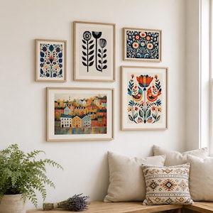 A set of framed art prints with folk art designs. The prints depict floral patterns, stylized birds, and a colourful townscape. The frames are light wood, and the art is displayed on a white wall.