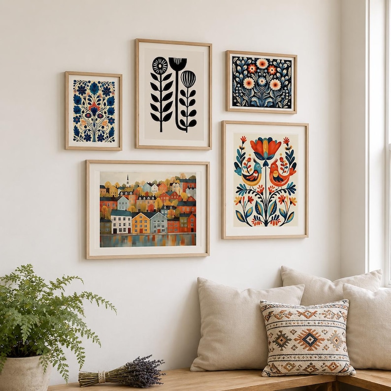 A set of framed art prints with folk art designs. The prints depict floral patterns, stylized birds, and a colourful townscape. The frames are light wood, and the art is displayed on a white wall.