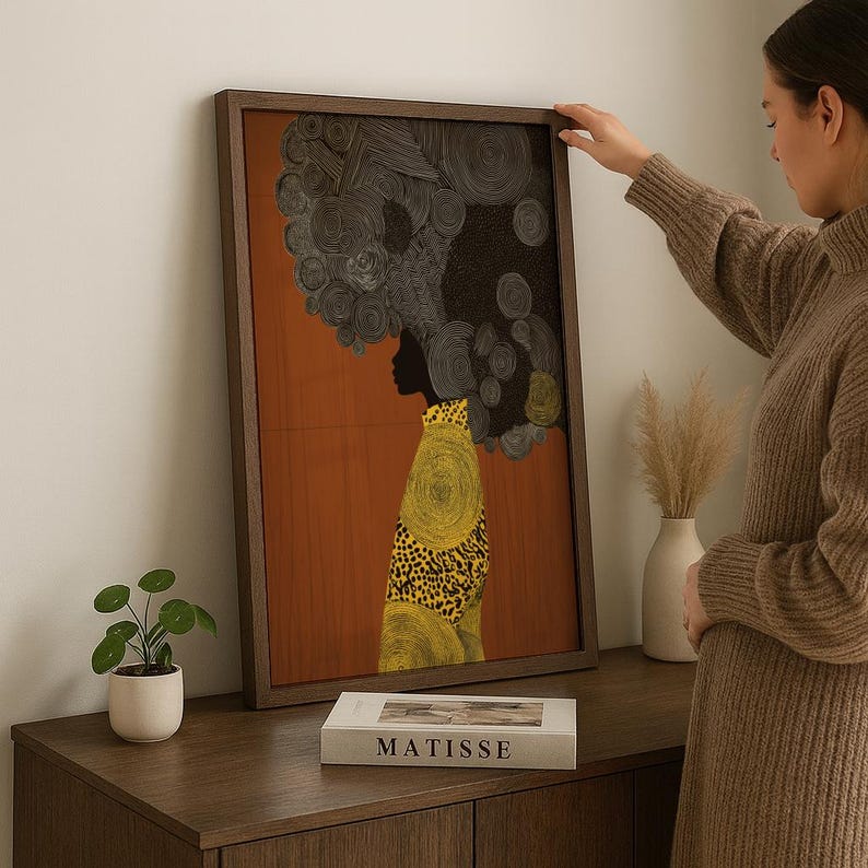Pu&ograve; includere: Una stampa artistica incorniciata raffigurante la silhouette di una persona con un'acconciatura grande e strutturata. La figura indossa un capo con motivi gialli e neri su sfondo arancione. L'opera d'arte &egrave; esposta su un mobile in legno.
