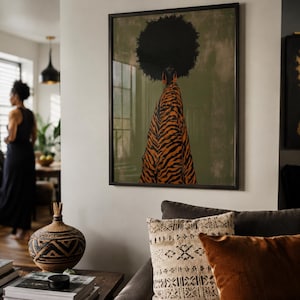 May include: A framed wall art print featuring a figure with a large afro hairstyle seen from behind, wearing a flowing orange and black tiger-striped garment against a muted olive green background, displayed in a living room setting.