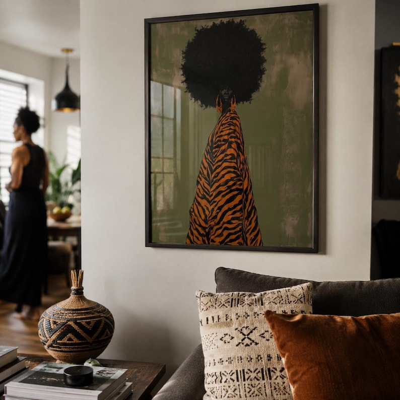 May include: A framed wall art print featuring a figure with a large afro hairstyle seen from behind, wearing a flowing orange and black tiger-striped garment against a muted olive green background, displayed in a living room setting.