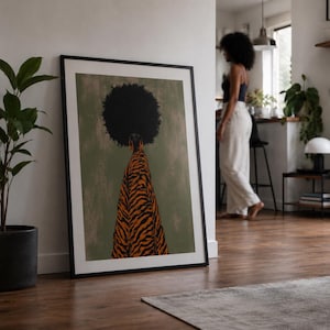 May include: A framed wall art print featuring a stylized illustration of a person with a large afro hairstyle wearing a flowing orange and black tiger-striped garment against a muted olive green background, displayed in a black frame.