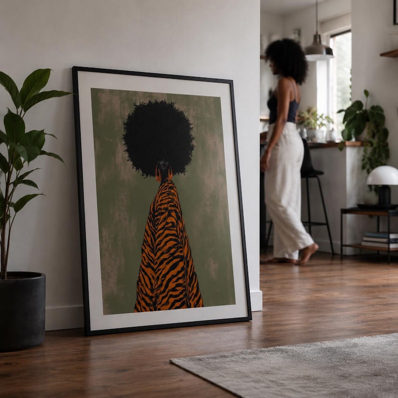 May include: A framed wall art print featuring a stylized illustration of a person with a large afro hairstyle wearing a flowing orange and black tiger-striped garment against a muted olive green background, displayed in a black frame.