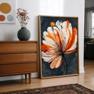May include: A framed art print depicting a large, stylized flower in white, orange, and peach hues against a dark blue backdrop. The artwork is displayed on a wooden floor. A wooden chest of drawers and vase are also visible in the room.