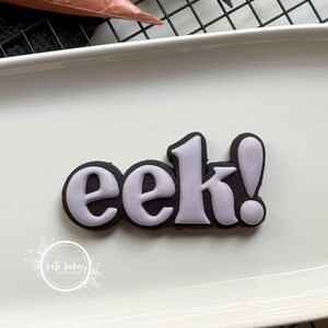 May include: A black cookie shaped like the word eek! with light purple icing lettering. The cookie is placed on a white rectangular tray. A circular logo with the text Kate Bakes Colorado is visible in the lower left corner of the image.