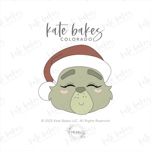 May include: A cartoon illustration of a green creature's face wearing a red and white Santa hat. The creature has closed eyes, pink cheeks, and a small smile. The text "kate bakes COLORADO" is at the top.