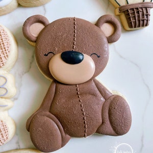 May include: A brown teddy bear cookie decorated with stitched details and a black nose. The cookie is sitting on a white marble surface.