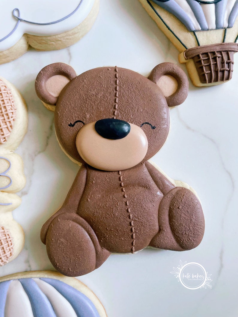 May include: A brown teddy bear cookie decorated with stitched details and a black nose. The cookie is sitting on a white marble surface.
