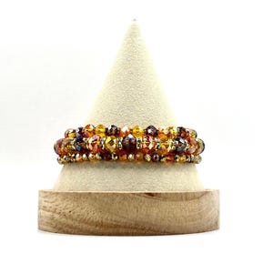 May include: A multi-strand bracelet with amber, gold, and brown glass beads. The bracelet is displayed on a wooden stand.
