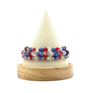 May include: A multi-strand bracelet with red, white, and blue crystal beads. The bracelet is displayed on a wooden stand.