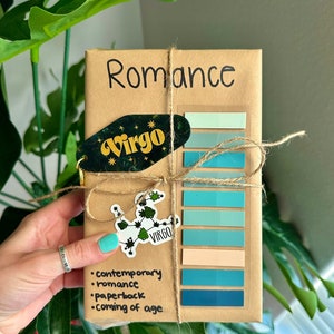 May include: A brown paper package wrapped with twine and decorated with stickers. The package is labeled "Romance" and has a sticker with the zodiac sign Virgo. The package also has a list of genres: contemporary, romance, paperback, coming of age.