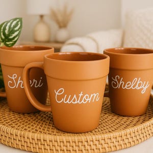 Op de afbeelding: Drie terracotta-kleurige keramische mokken met handvatten, elk met een naam in witte letters: "Sheri", "Custom" en "Shelby". De mokken zijn gerangschikt op een ronde geweven dienblad, met een groene plant zichtbaar.