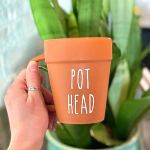 May include: A terracotta-colored ceramic mug with a white handle. The mug has the words "POT HEAD" written on it in white.