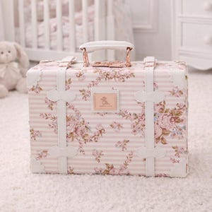 May include: A decorative, floral-patterned suitcase with pink and white stripes. The suitcase features a white handle, white straps, and a small rectangular emblem. The background includes a white crib and a stuffed bunny.