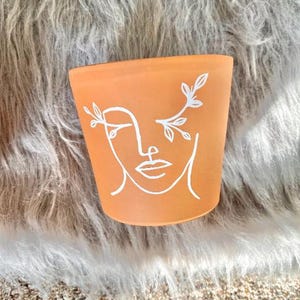 Puede incluir: Una maceta de color terracota con un dibujo lineal blanco de una cara. La cara tiene un ojo cerrado y está adornada con diseños blancos en forma de hojas. La maceta es cilíndrica y se asienta sobre una superficie suave y clara.