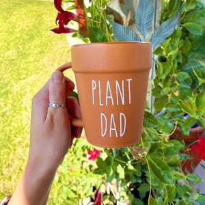 May include: A terracotta-colored ceramic mug with a white handle and the words "Plant Dad" written on the front. The mug is being held by a hand wearing a silver ring.