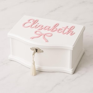 Pode incluir: Uma caixa de joias de madeira branca com uma parte superior curva e fechadura com chave. O nome "Elizabeth" está escrito em letras rosa na tampa, com um laço rosa. Uma borla branca pende da chave.