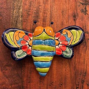 May include: A colorful ceramic bee with blue, green, yellow, orange, and red accents. The bee has wings with floral designs and a striped body.
