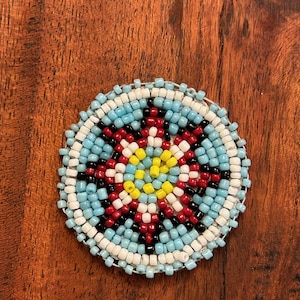May include: A round beaded design with a blue, white, red, black and yellow colour scheme. The beads are arranged in a circular pattern, creating a geometric design.
