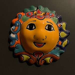 Puede incluir: Un sol de cerámica colorido con una cara sonriente, pintado en azules, verdes, amarillos y rojos brillantes. El sol tiene un diseño floral alrededor de los bordes.