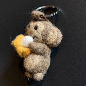 Op de afbeelding: Een bruine en witte vilten aap sleutelhanger die een gele vilten banaan vasthoudt.