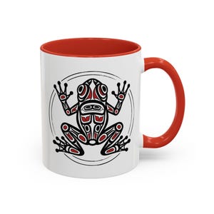 May include: White ceramic mug with a red interior and handle. The mug features a black, red, and white frog design within a circular frame. The frog design is a stylized, symmetrical illustration.