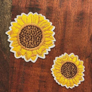 Puede incluir: Dos parches bordados de girasol con pétalos de color amarillo brillante y un centro marrón oscuro texturizado. Cada parche tiene un borde blanco y un contorno festoneado, con costuras detalladas que imitan la apariencia natural de la flor.