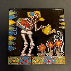 May include: Square ceramic tile depicting a skeleton watering flowers. The skeleton wears a yellow hat and holds a yellow watering can. The tile has a black background with colourful floral and geometric border designs.