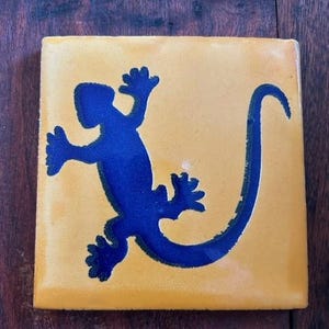 May include: A yellow ceramic tile with a blue silhouette of a lizard with a curled tail.
