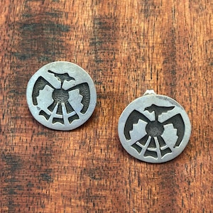May include: Two round, silver-toned earrings with a Native American-inspired design. Each earring features a central sun-like motif surrounded by a stylized bird or eagle shape. The earrings are displayed on a wooden surface.