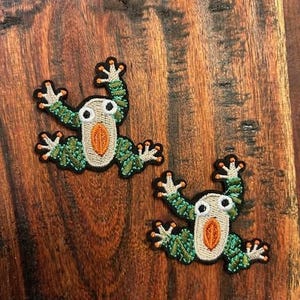May include: Two green and orange embroidered frog patches on a brown wooden surface.