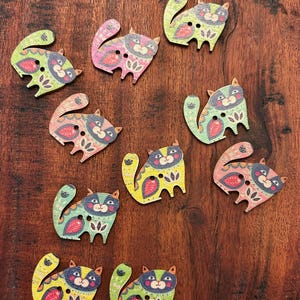 Könnte beinhalten: Eine Sammlung von Holzknöpfen in Katzenform in verschiedenen Farben wie Gelb, Rosa, Grün und Blau. Jeder Knopf hat ein einzigartiges dekoratives Design und zwei kleine Löcher zum Annähen. Die Knöpfe liegen auf einer dunklen Holzoberfläche.