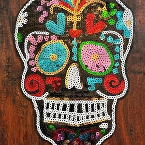May include: A colorful sequined sugar skull patch with black, white, red, blue, green, yellow, and pink sequins. The skull has a floral design and is perfect for adding a festive touch to clothing, bags, or other crafts.