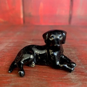 May include: A small, glossy black figurine of a dog in a resting pose. The dog is lying down with its front legs extended and its head raised slightly. The figurine is on a red surface.