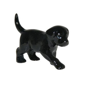 May include: A small, glossy black Labrador puppy figurine. The dog is depicted in a walking pose with its tail raised. The figurine has blue eyes and is set against a white background.