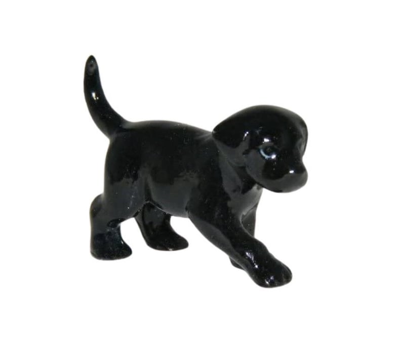 May include: A small, glossy black Labrador puppy figurine. The dog is depicted in a walking pose with its tail raised. The figurine has blue eyes and is set against a white background.