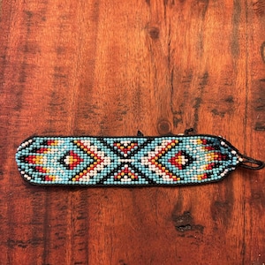 May include: A beaded bracelet with a geometric pattern in turquoise, red, yellow, white, and black. The bracelet is rectangular with a black backing and a black string closure. The bracelet is resting on a wooden surface.