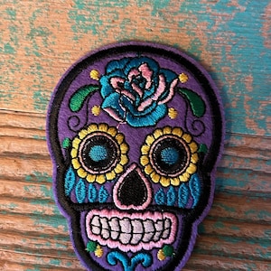 May include: Purple embroidered sugar skull patch with a blue and pink flower, yellow and blue eyes, and a black nose. The skull is surrounded by a black outline.