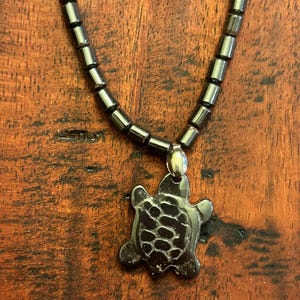 May include: A dark grey beaded necklace with a turtle pendant. The turtle pendant has a textured design. The necklace is displayed on a wooden surface.