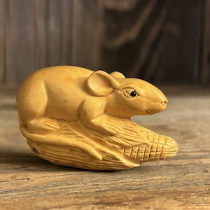 Puede incluir: Una pequeña talla de madera de un ratón sentado sobre un montón de maíz. El ratón es de color marrón claro y tiene ojos negros. El maíz también es de color marrón claro y tiene una talla detallada.