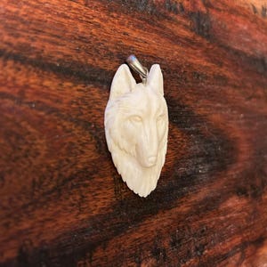Könnte beinhalten: Ein weißer Wolf-Anhänger aus Knochen mit einer silbernen Öse. Der Anhänger ist detailliert mit realistischen Merkmalen und ist ein wunderschönes Beispiel für Tierkunst.
