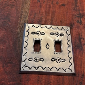 May include: Silver-toned metal light switch cover with a decorative border of small dots and oval shapes. The cover has two rectangular openings for switches and a diamond-shaped detail in the center.